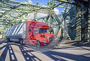 Long haul bright red big rig semi truck transporting cargo in dry van semi trailer running on the arched truss bridge