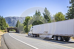 Long haul white big rig semi truck with dry van semi trailer delivering cargo running on the winding road with forest and mountain