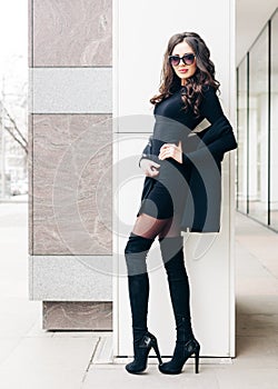Long-legged beauty of a brunette in sunglasses posing. A small black outfit, high boots with high heels. Fashion. Style.