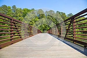 A long metal rust colored bridge over a lake surrounded by lush green trees and plants with blue sky at Cauble Park