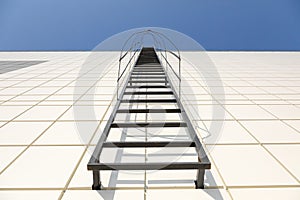 Long metal staircase on the gray modern facade of an industrial building, warehouse or shopping center against the blue sky. Fire