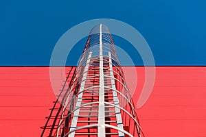 Long metal staircase on the red modern facade of an industrial building, warehouse or shopping center against the blue sky. Symbol