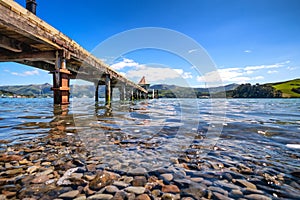 A long pier at Akaroa, New Zealand. The water is so clear that one can see pebbles under the water. There are rolling hills in the