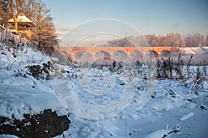 Long red brick bridge over Venta river in misty, sunny, snowy winter morning with frosted trees, Kuldiga, Latvia