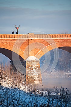 Long red brick bridge over Venta river in misty, sunny, snowy winter morning with frosted trees, Kuldiga, Latvia