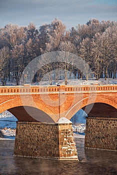 Long red brick bridge over Venta river in misty, sunny, snowy winter morning with frosted trees, Kuldiga, Latvia