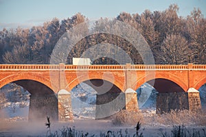 Long red brick bridge over Venta river in misty, sunny, snowy winter morning with frosted trees, Kuldiga, Latvia