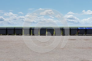 Long row of black garbage containers outside under the blue sky with white clouds