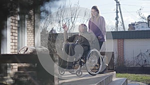 Long shot of adult female invalid tender talking with old Caucasian man and rolling wheelchair away. Smiling nurse