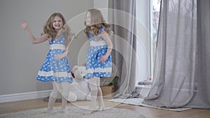 Long shot portrait of elegant twin sisters dancing and spinning indoors. Cheerful Caucasian girls resting at home. Joy