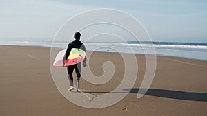 Long shot of surfer with artificial leg walking along beach
