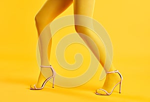 Long slim legs of young woman in yellow tights and stylish high heels shoes posing on yellow background