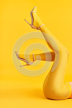 Long slim legs of young woman in yellow tights and stylish high heels shoes on yellow background.