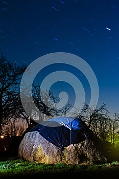 Long star tracks in clear deep blue night sky, beautiful countryside landscape with a haycock in a farm garden