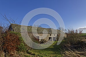 A long Stone built barn with a pitched slate roof, abandoned and set within the overgrown grounds of a Farmhouse