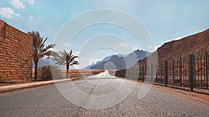 Long straight road with desert hills in distance, brick wall and iron fence on sides. Scenery at the entrance to Wadi