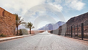 Long straight road with desert hills in distance, brick wall and iron fence on sides. Scenery at the entrance to Wadi Rum