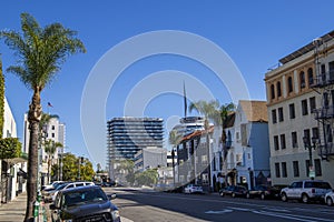 A long street lined with parked cars, office buildings, apartments, lush green palm trees and shops and cars driving