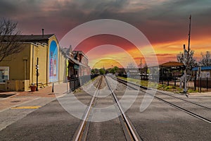 A long stretch of railroad tracks covered in gravel with a black metal fence and buildings along the tracks and blue sky