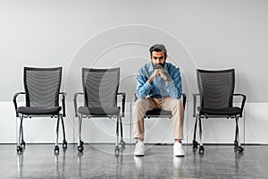 Long time of waiting. Thoughtful serious indian businessman holding hands on chin sitting on chair against grey wall