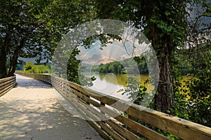 A long winding boardwalk along the river with a wooden rail along the sides with vast still river water, lush green trees