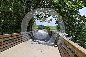 A long winding boardwalk along the river with a wooden rail along the sides with vast still river water, lush green trees