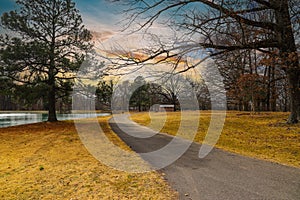 A long winding footpath along side of a green lake in the park surrounded by yellow winter grass, bare winter trees