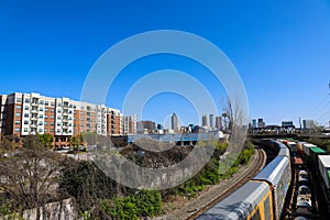 A long winding set of railroad tracks and a train surrounded by lush green and bare winter trees with skyscrapers