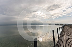 Long wooden boardwalk pier over water with a mountain landscape under an overcast sky