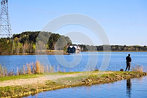 A long wooden dock on the lake with vast blue lake water and an African American man fishing on the banks of the lake