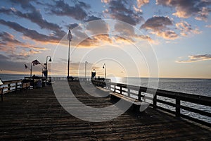 A long wooden pier with curved light posts along the pier with vast miles of blue ocean water and blue sky and powerful clouds