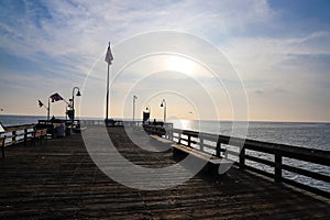 A long wooden pier with curved light posts along the pier with vast miles of blue ocean water and blue sky and powerful clouds