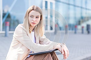 Longing young woman sitting alone on street sidewalk on urban background modern building. Sad girl Student or businesswoman misses