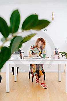 Longshot of a focused artist woman painting behind work desk shot through plant