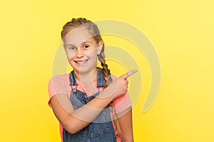 Look, advertise here! Portrait of cheerful cute little girl in denim overalls pointing to side copy space