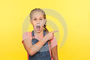 Look, crazy advertise! Portrait of amazed cute little girl in denim overalls pointing to side copy space