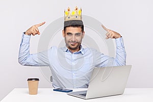 Look, I`m king! Proud authoritative businessman pointing at crown on head while sitting in office workplace