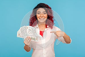 Look, I`m rich! Portrait of successful happy woman with fancy red hair pointing to dollar bills