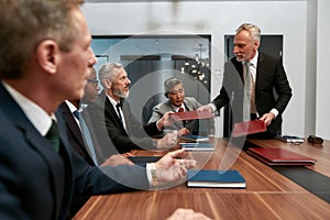 Look please. Mature businessman in formal wear giving folders to his multicultural colleagues while having a meeting in