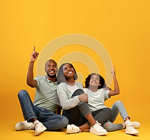 Look there. Happy african american family pointing fingers up at empty space, sitting on floor over yellow background