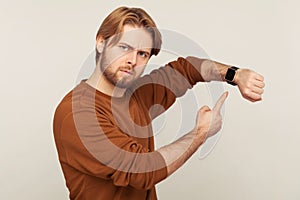 Look at time! Portrait of angry impatient man with beard in sweatshirt pointing wrist watch and looking annoyed