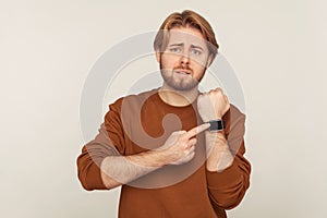 Look at time! Portrait of worried impatient man with beard in sweatshirt pointing wrist watch and looking anxious