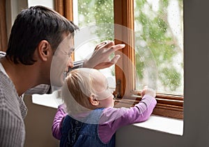 Look at what a beautiful day it is outside. A young father and his baby girl looking at something beyond the window.