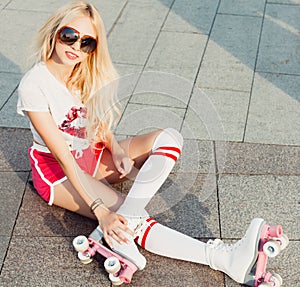 A looker leggy long-haired young blonde woman in a vintage roller skates, sunglasses, T-shirt shorts sitting on road. Summer portr