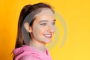 Looking away. Portrait of young beautiful girl posing in pink hoodie over yellow studio background. Concept of youth