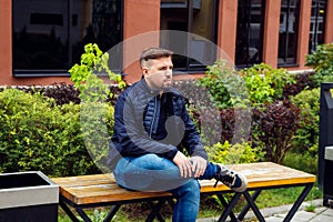 Looking at the camera young Caucasian businessman 34 years old man with goatee beard sits on bench in modern landscaped garden in