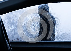 Looking through a car window at a mysterious scary hooded figure standing outside. On a rainy winters day