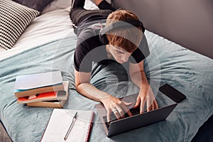 Looking Down On Male College Student Wearing Headphones Lying On Bed Working On Laptop