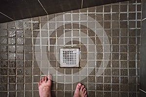 Looking down to hotel room shower drain grid, wet tiles on floor, man feet visible in lower part