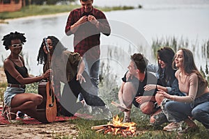 Looking at guitar. Group of people have picnic on the beach. Friends have fun at weekend time